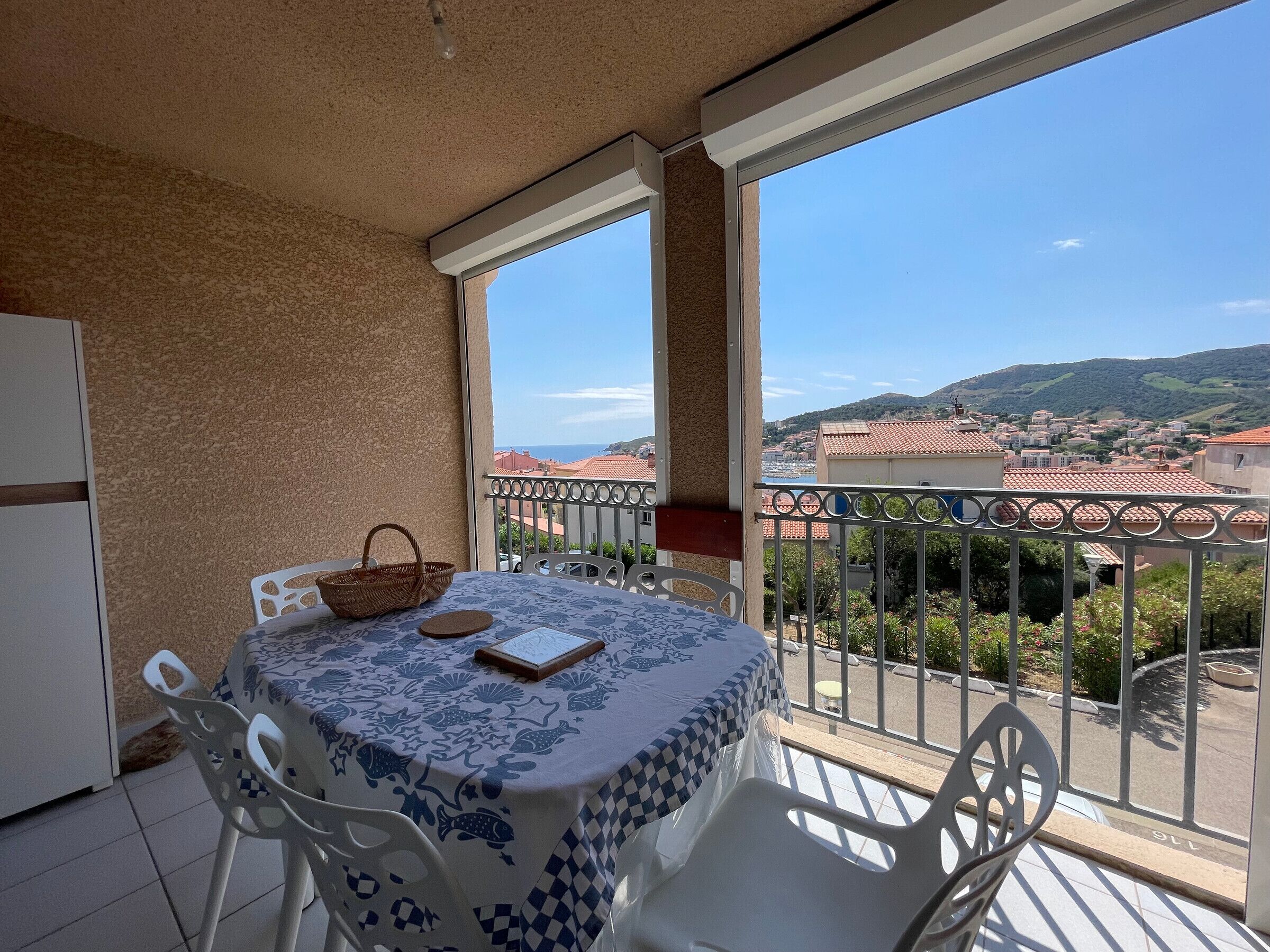 A balcony with a table set for two, a basket, and a book. The balcony overlooks a town with red-roofed buildings and a view of the sea.