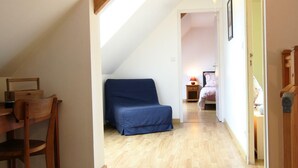 A narrow hallway with a wooden chair, a blue sofa, and a bed in the background. - Bright house with fireplace and enclosed garden (Le Plessis-Lastelle)