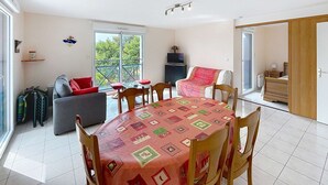 Un comedor con una mesa cubierta por un mantel estampado, rodeada de sillas de madera. Un sofá gris y una silla roja están ubicados contra la pared, con un televisor en un mueble cercano. Una puerta corredera da acceso a un dormitorio con una cama de madera y una mesita de noche.