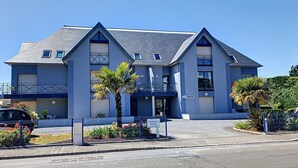 Un edificio moderno de dos pisos con fachada azul, entrada protegida por un portón y un pequeño jardín con palmeras.