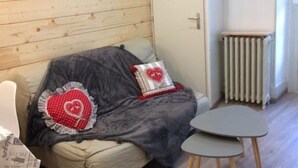 A cozy room with a wooden wall, a sofa with a grey blanket, two heart-patterned pillows, and a small table. - T2 Apartment Cauterets Center - 4 People, Wifi (Cauterets)