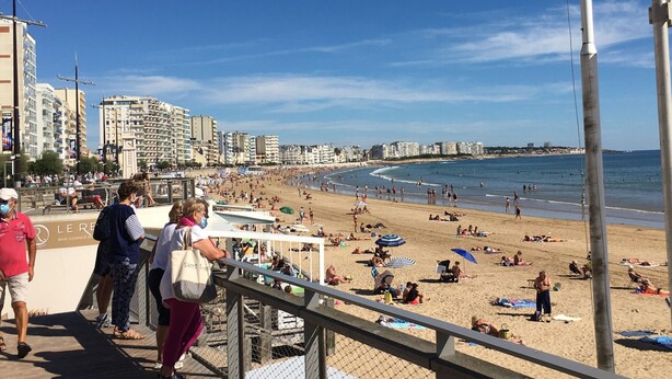 A beach with people sunbathing and swimming, a bar lounge, and buildings in the background.