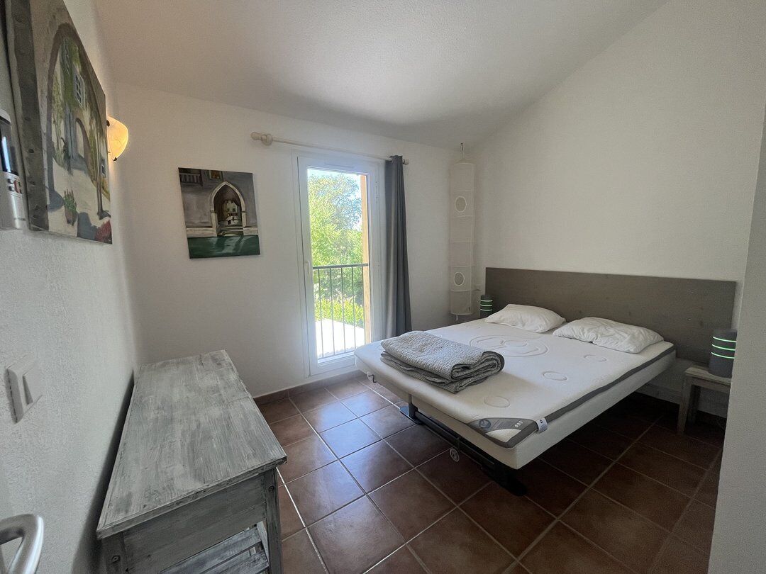 A bedroom with a bed, a small bedside table, a nightstand, a window with curtains, and a wall-mounted shelf.