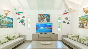 Un saló modern amb un televisor de pantalla plana gran, dos sofàs amb coixins tropicals, una taula de menjador i diverses obres d'art a la paret, incloent-hi peixos, vaixells i paisatges pintats.