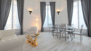 Un soggiorno moderno con un divano bianco, un tavolino da caffè in vetro con base decorativa, un tavolo da pranzo con sedie e tende grigie.