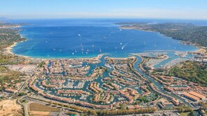 Vue aérienne d’une ville côtière avec un port de plaisance rempli de bateaux, entourée de bâtiments résidentiels et surplombée d’un ciel bleu clair.