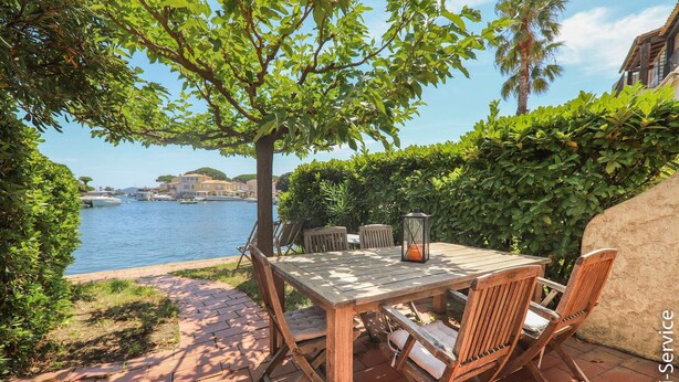 Une terrasse extérieure avec une table et des chaises en bois, une lanterne, et une vue sur l’eau et les bâtiments en arrière-plan.