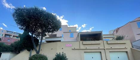 Un immeuble résidentiel à la façade beige, avec un arbre devant et un ciel bleu clair.