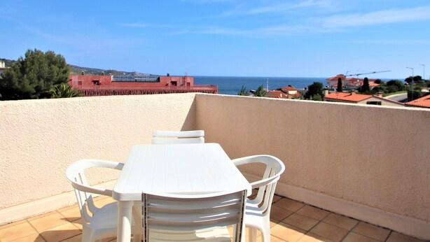 A balcony with a white table and chairs, overlooking a coastal area with buildings and a clear sky.