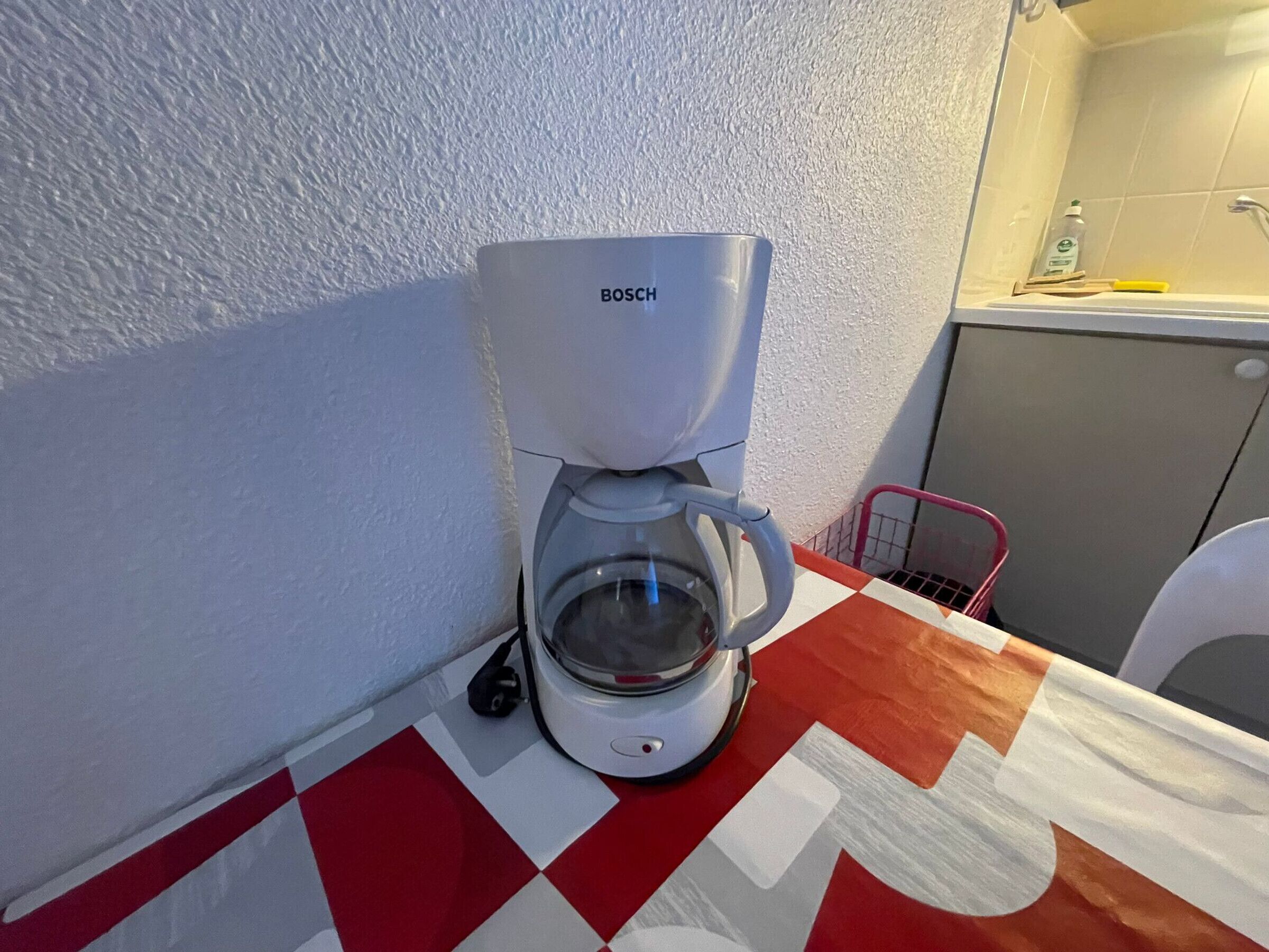 Une machine à café Bosch est posée sur une table à carreaux dans une salle de bain équipée d’un lavabo et d’un porte-serviettes.
