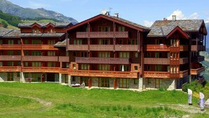 A multi-story wooden hotel with balconies and a stone foundation, set against a mountainous backdrop. - 3-room apartment, 6 pers, balcony, pets allowed, optional parking (Valmorel)