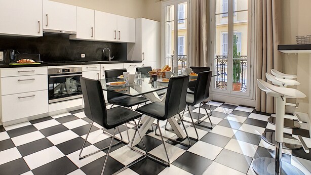 Une cuisine moderne avec des meubles blancs, un plan de travail noir et un sol à carreaux. On y trouve une table à manger avec des chaises et une fenêtre avec des rideaux.