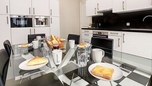 Une cuisine moderne avec des meubles blancs, un plan de travail noir et une table à manger en verre, mise avec des assiettes, des verres et un panier de pain.