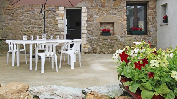 Un patio al aire libre con un edificio de piedra, sillas de plástico blanco, una mesa y un paraguas grande.