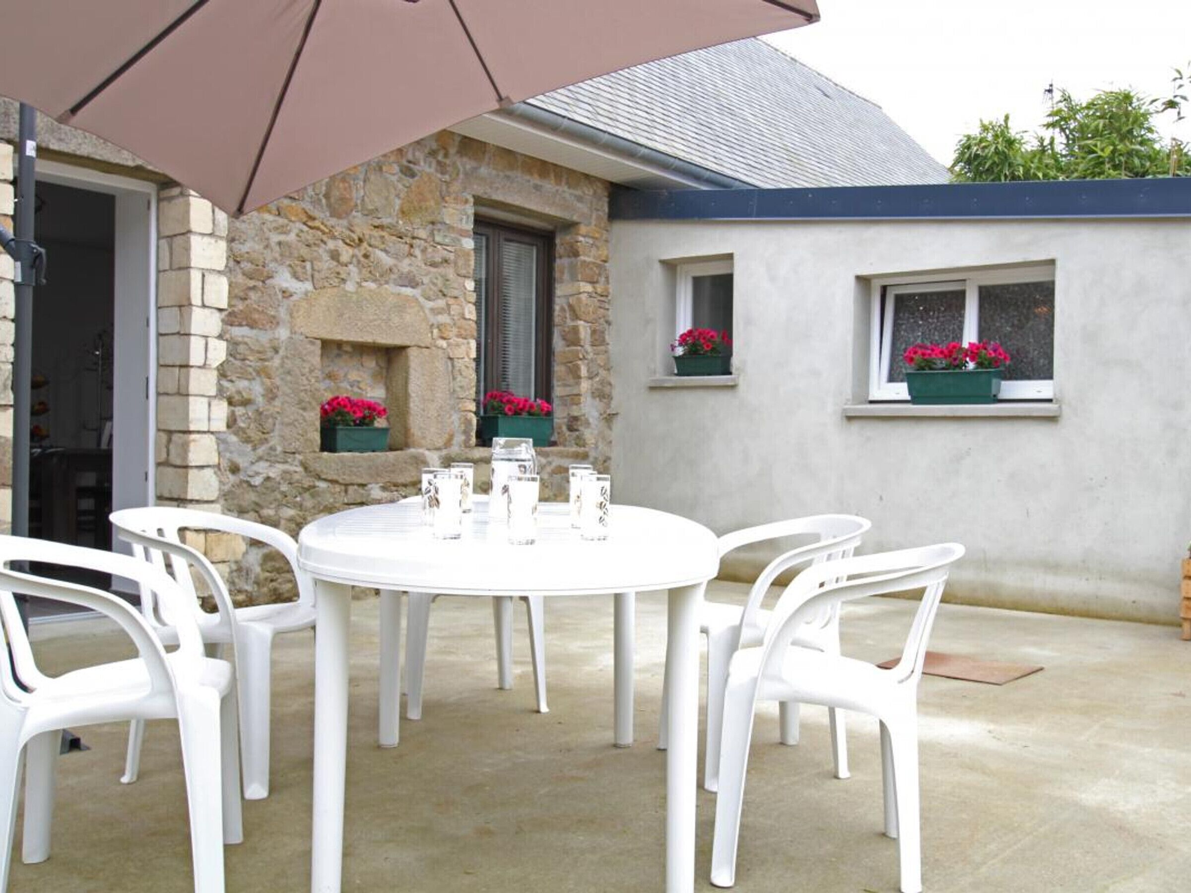 Une table ronde entourée de chaises blanches, protégée par un grand parasol, se trouve dans un patio extérieur dont les murs sont en pierre et les fenêtres sont ornées de jardinières fleuries.