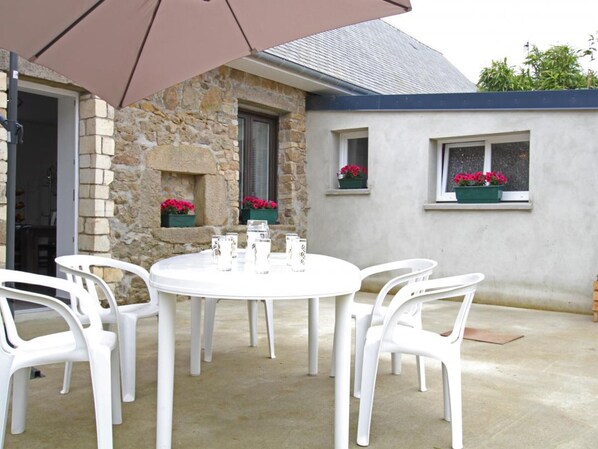 Una mesa redonda con sillas blancas y un paraguas grande en un patio al aire libre con muros de piedra y ventanas con jardineras.