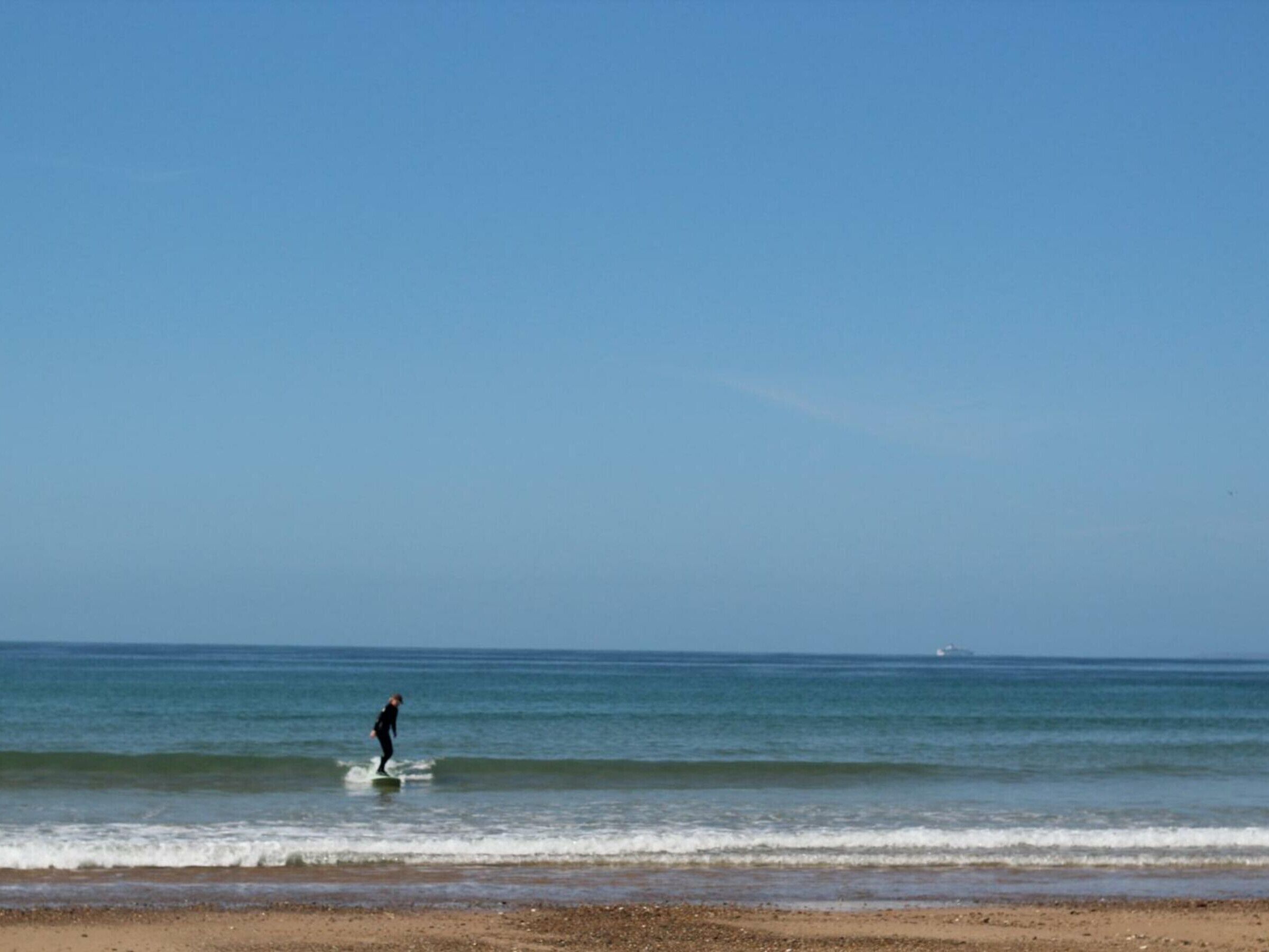 A beach with a person surfing, clear skies, and calm sea.
