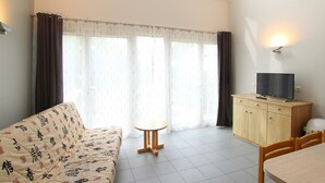 Uma sala de estar moderna com um sofá, uma mesa redonda pequena, uma TV sobre um móvel de madeira e janelas amplas com cortinas.