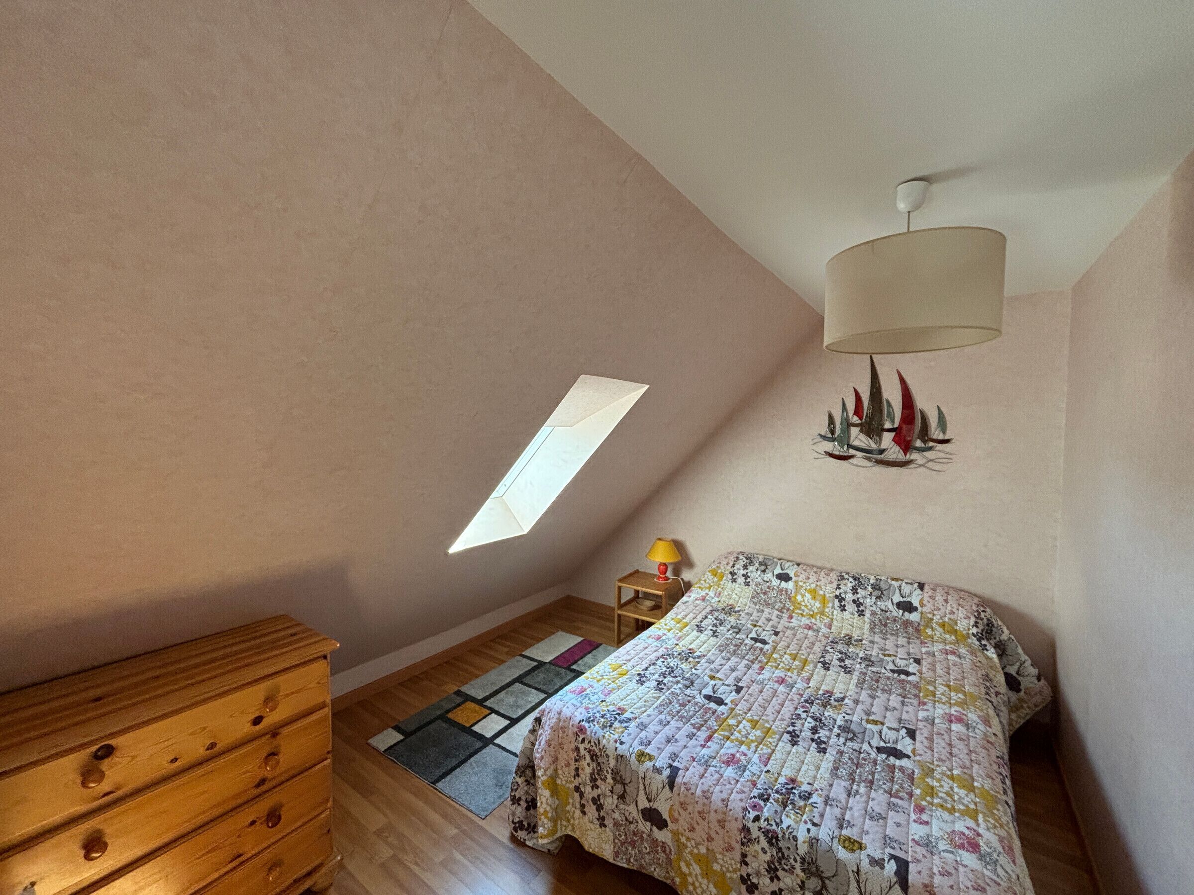 Un dormitorio con techo inclinado, una cama con colcha floral, un aparador de madera, una mesita con lámpara y una decoración de velero montado en la pared.