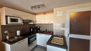 A compact kitchen with a microwave, stove, sink, and a clock on the wall. - 2-Bedroom Apartment - 50m from the beach, parking (Les Sables-d'Olonne)