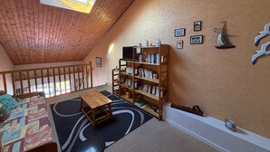 A cozy attic room with a sloped ceiling, a skylight, a wooden bookshelf, a small table, and a bed with a patterned bedspread. - 2-Bedroom Apartment - 50m from the beach, parking (Les Sables-d'Olonne)