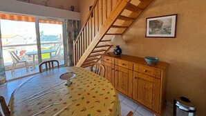 A dining area with a table covered in a fruit-patterned tablecloth, wooden cabinets, a staircase, and a view through a glass door. - 2-Bedroom Apartment - 50m from the beach, parking (Les Sables-d'Olonne)