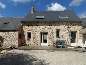 A stone building with a sloped roof, two windows with shutters, a wooden deck, and outdoor seating.