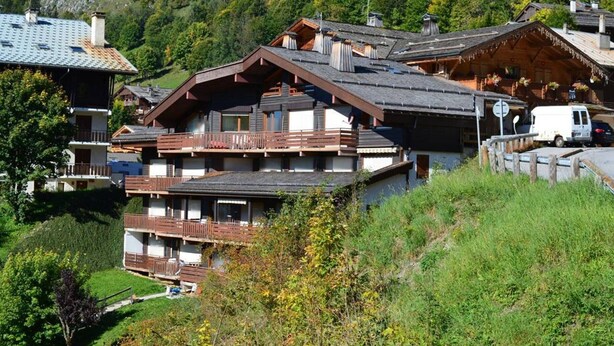A multi-story chalet with wooden balconies, set in a mountainous area with lush greenery.