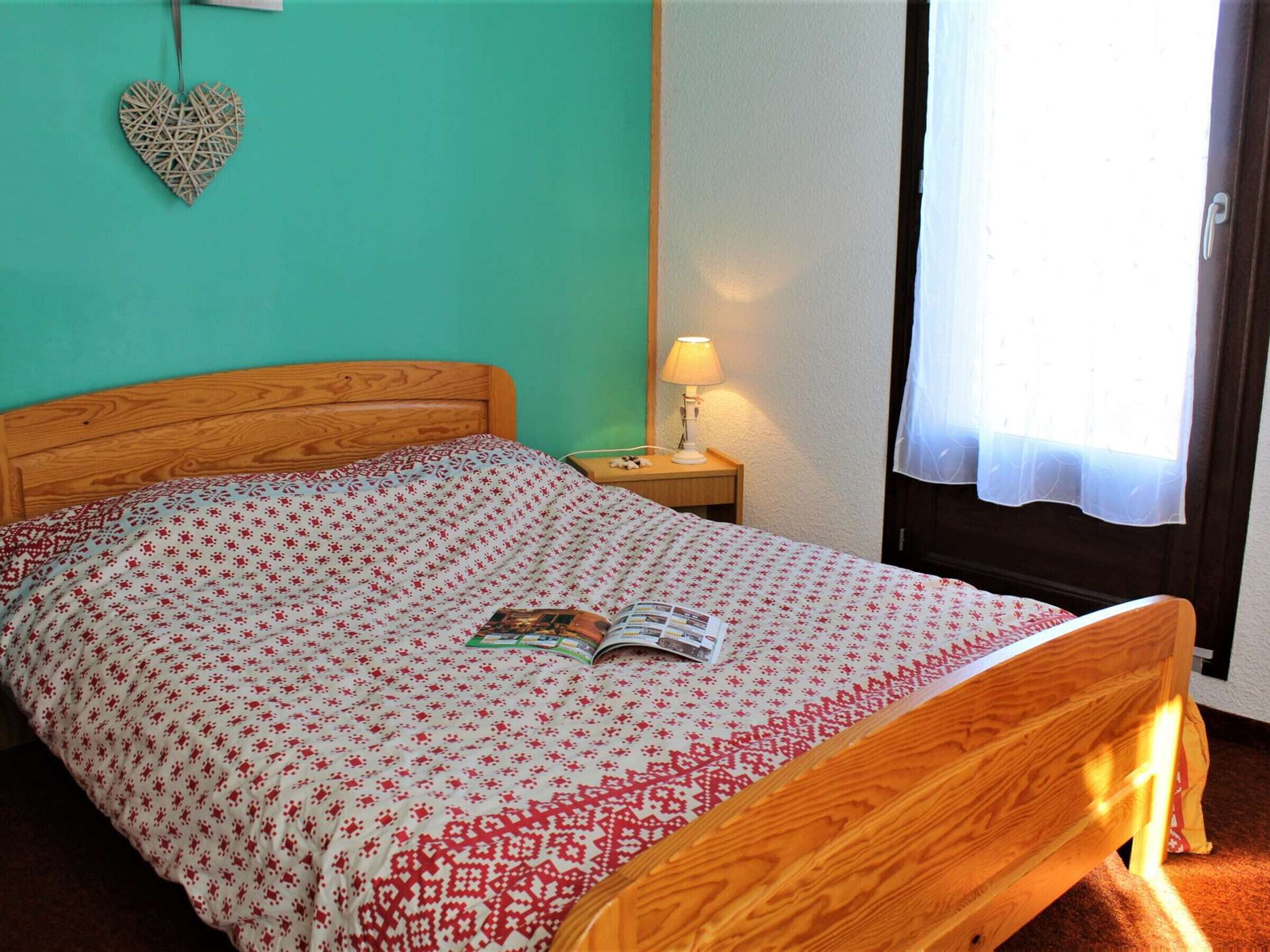 A bed with a wooden headboard, a patterned bedspread, a heart-shaped decoration on the wall, a bedside lamp, and a window with a curtain.