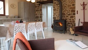 A cozy living room with a stone wall, a fireplace, white furniture, and a dining area with a table and chairs. - Charming cottage with Internet near Mont Saint-Michel (Les Chambres-Le Grippon)