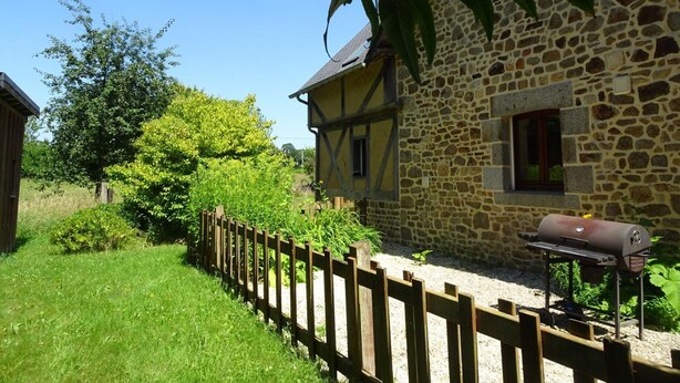 A stone house with a wooden fence, a barbecue grill, and lush greenery. - Charming cottage with Internet near Mont Saint-Michel (Les Chambres-Le Grippon)