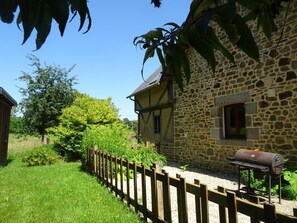 A stone house with a wooden fence, a barbecue grill, and lush greenery. - Charming cottage with Internet near Mont Saint-Michel (Les Chambres-Le Grippon)