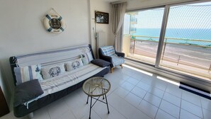 A room with a sofa, a small round table, a 'WELCOME ABOARD' sign, a view of the sea through a large window, and a balcony with a net.