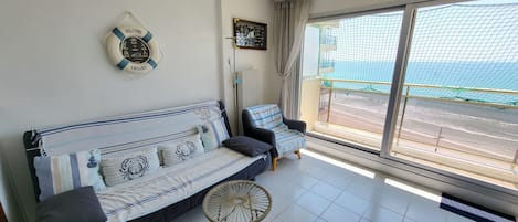 A room with a sofa, a small round table, a 'WELCOME ABOARD' sign, a view of the sea through a large window, and a balcony with a net.