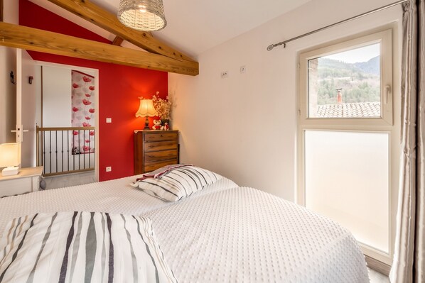 Ein Schlafzimmer mit Bett, Kommode, Nachttisch, Lampe und einem Fenster mit Blick ins Freie.