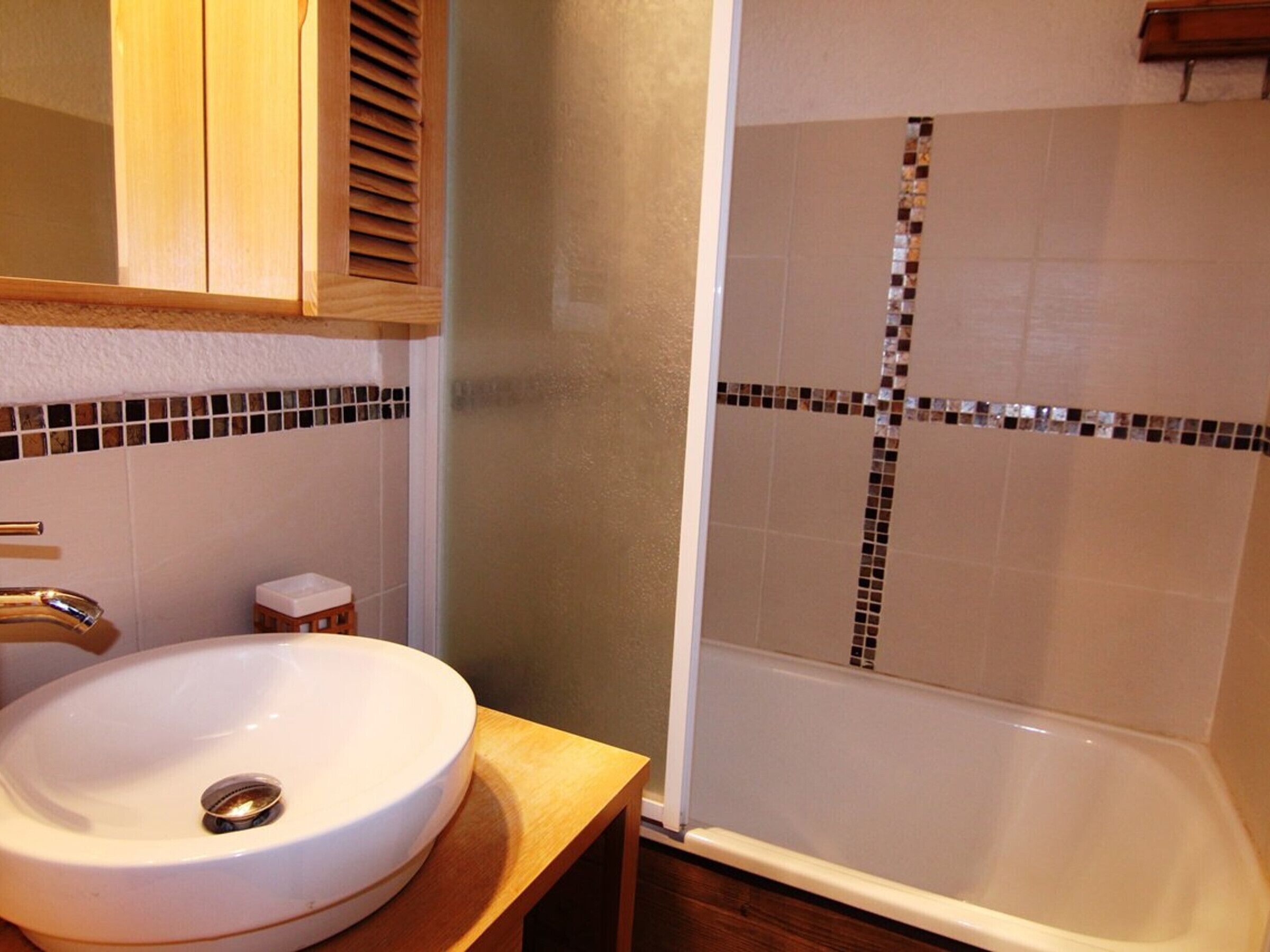 A bathroom with a white sink, a wooden cabinet above it, and a shower with mosaic tiles.