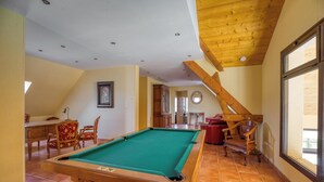A pool table in a room with wooden beams, a dining area, and a view through large windows.