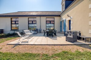 Eine Terrasse mit Gartenmöbeln, darunter Stühle und ein Tisch, sowie ein Gebäude mit großen Fenstern und einer blauen Tür.
