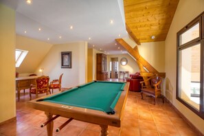 Ein Billardtisch in einem Raum mit schräg verlaufender Decke, Holzbalken und einem Essbereich mit roten Stühlen.