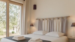 Un letto rifatto con lenzuola bianche e una coperta scura, due lampade ai comodini e vista sull'esterno attraverso ampie finestre.