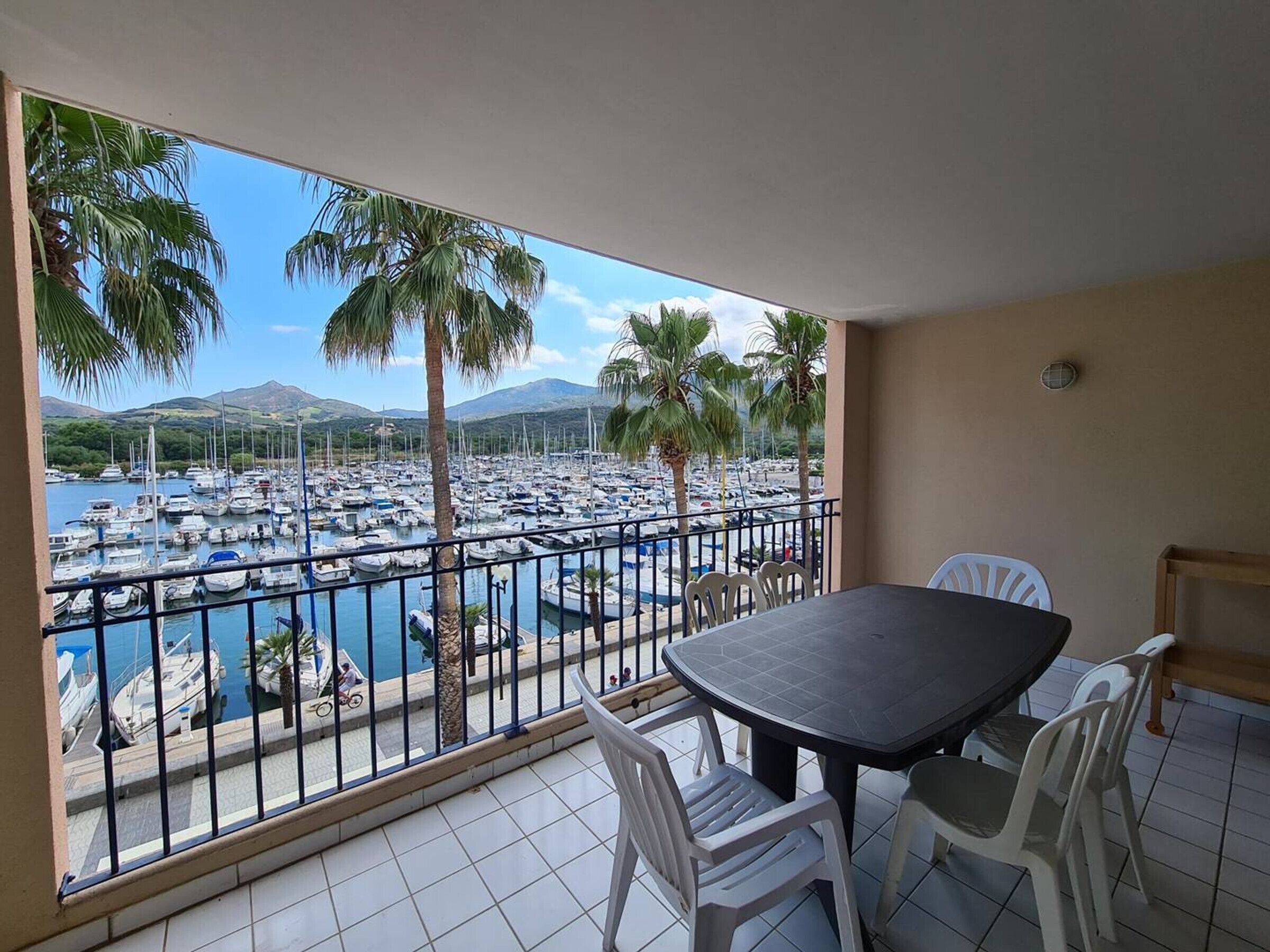 A balcony with a view of a marina and palm trees, a round table with chairs, and a tiled floor.