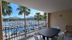 A balcony with a view of a marina and palm trees, a round table with chairs, and a tiled floor.