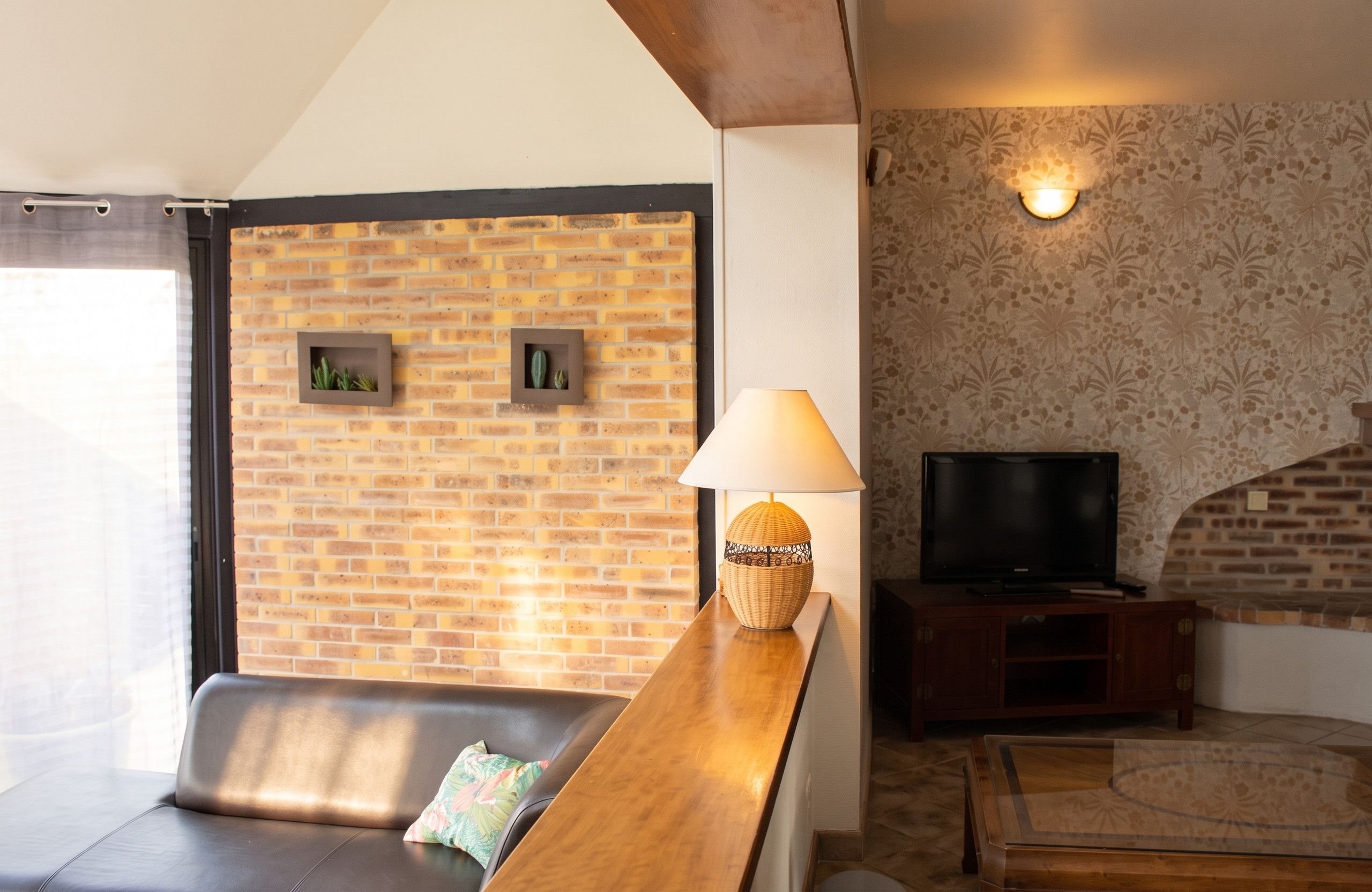 A living room with a brick wall, a sofa, a wooden table with a lamp, a television, and a patterned wall.