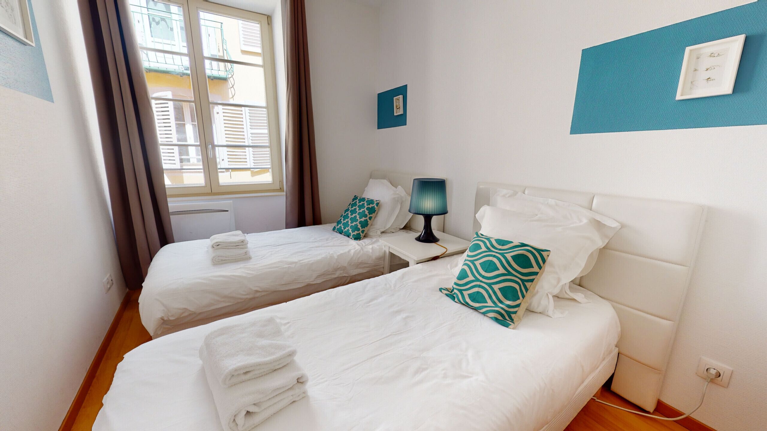 A hotel room with two beds, a window with blinds, a bedside table with a lamp, and a wall-mounted power outlet.