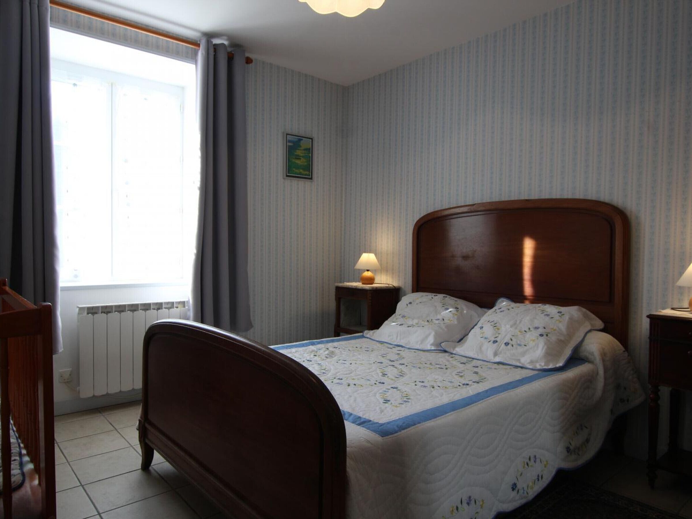A bedroom with a wooden bed, a radiator, a window with curtains, and a small bedside table with a lamp.
