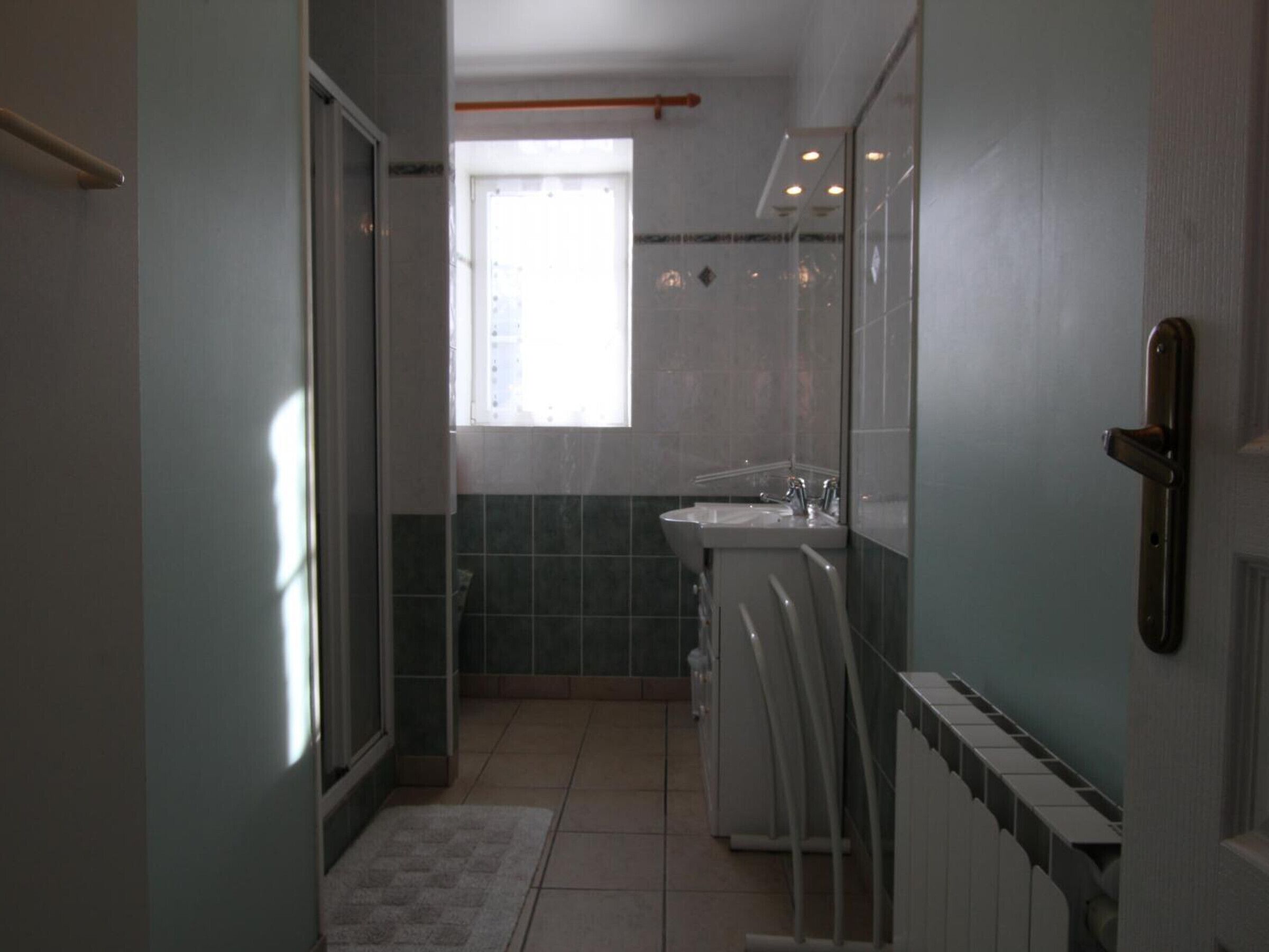 Een smalle gang die uitkomt bij een badkamer met een raam, een wastafel en een radiator.