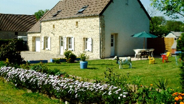 A stone house with a sloped roof, a small garden with flowers, and a table with chairs outside.