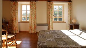 Ein Schlafzimmer mit Holzbalken, zwei Fenstern mit Blumenvorhängen, einem Bett mit gemustertem Überwurf, einem Sessel und einem Nachttisch mit Lampe.