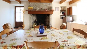 Ein Esszimmer mit einem runden Tisch, der mit einer Tierprint-Tischdecke bedeckt ist, umgeben von Holzstühlen. Auf dem Tisch steht eine blaue Vase, und im Hintergrund ist ein Steinkamin mit Kaminvorsatz zu sehen.