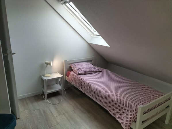 Ein kleines Zimmer mit einem Einzelbett, schräg verlaufender Decke und einem Dachfenster, ausgestattet mit einem Nachttisch, einer Lampe und einer Steckdose.
